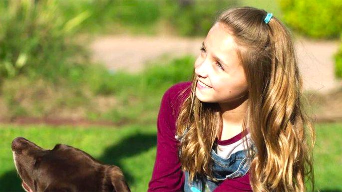 Menina loira olhando para o lado e sorrindo, com um cachorro castanho