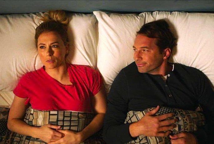 Casal deitado na cama; ela com expressão preocupada e ele olhando para ela.