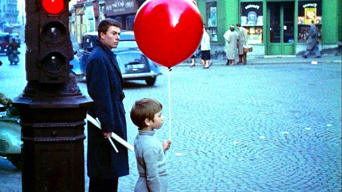 Garoto loiro na calçada, segurando um balão vermelho