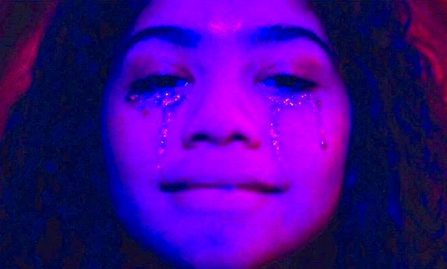 Imagem roxa, com Rue sorrindo, usando uma maquiagem de purpurina que simula lágrimas debaixo dos olhos