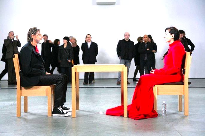 Mulher com vestido vermelho e homem de roupa escura sentados frente a frente