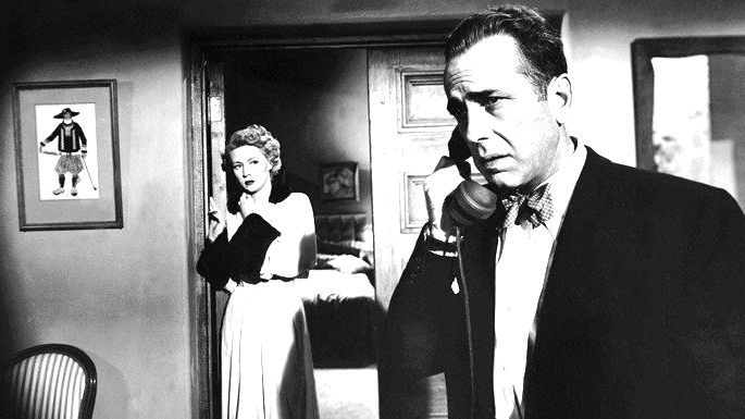 Imagem em preto e branco, com um homem falando no telefone e, no fundo, uma mulher escutando atrás da porta